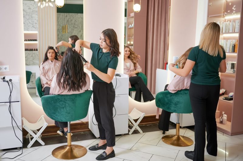 Young women sitting in chairs in front of mirrors while hairdressers styling clients hair. Two female hairstylists curling client hair and doing hairstyle in modern beauty salon.